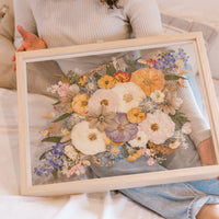Beautiful pressed florals displayed in a floating glass frame held by a bride admiring her bouquet preservation.