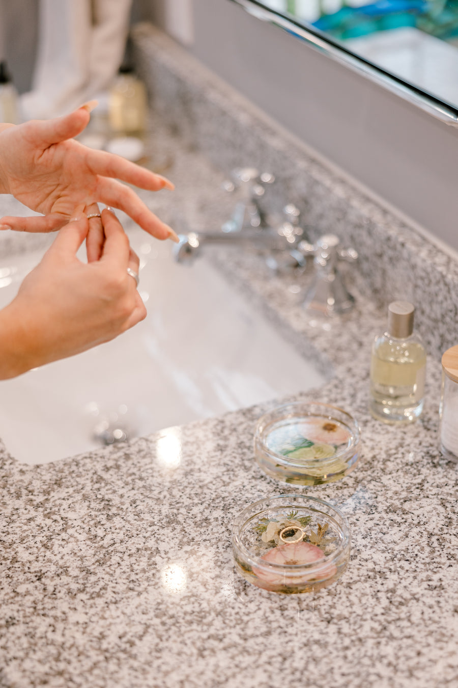 A woman removes her rings and sets them on top of a pressed flower coaster that sits beside her bathroom sink.