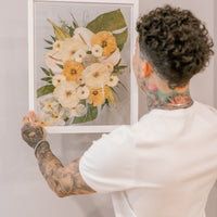 A groom hanging a bouquet preservation piece in his home that featured pressed tropical flowers such as preserved monstera leaves, pressed roses, and pressed orchids. 