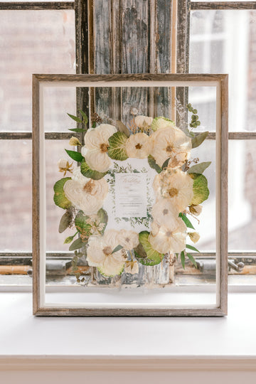 A beautiful bouquet preservation featuring white pressed roses and a preserved wedding invitation surrounded by pressed greenery in a barn wood frame.