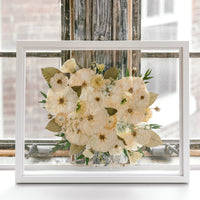 A beautiful pressed white bouquet with pops of greenery featured in a brightly lit window sill. Proudly designed by Element Preservation.
