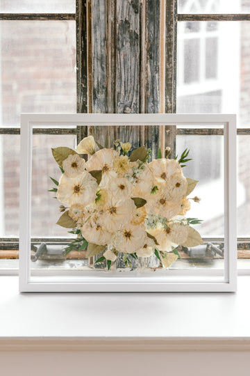 A beautiful pressed white bouquet with pops of greenery featured in a brightly lit window sill. Proudly designed by Element Preservation.