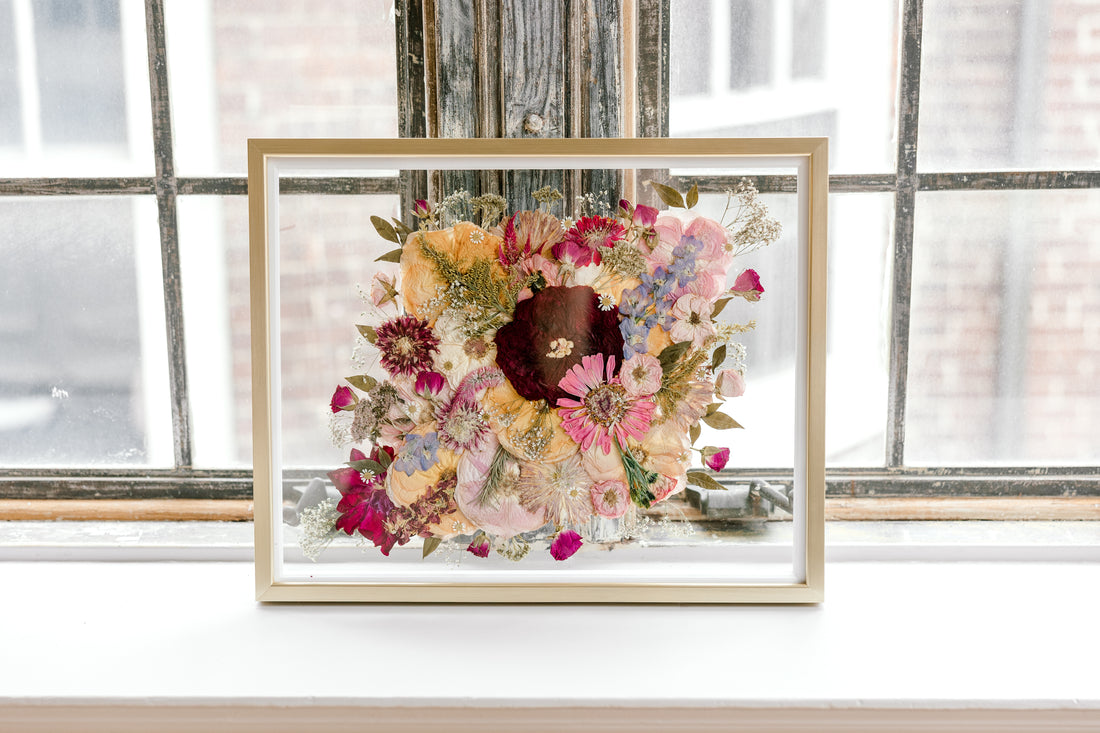 Brightly colored pressed florals are featured in this preserved bridal bouquet. The pressed flower frame is displayed in a gold wood frame against rustic windows.