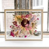 Brightly colored pressed florals are featured in this preserved bridal bouquet. The pressed flower frame is displayed in a gold wood frame against rustic windows.