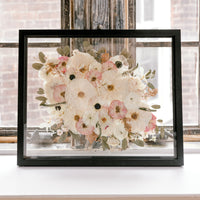 A pressed floral bouquet frame with white, pink, and green pressed flowers. Showcased in a black wood floating frame, this bouquet preservation shines beautifully in its forever home.