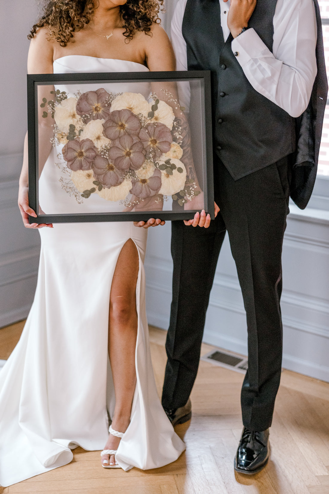 A happy couple stands holding their wedding bouquet preservation featuring pressed white roses and pressed purple roses with accents of pressed baby's breath and pressed eucalyptus. 