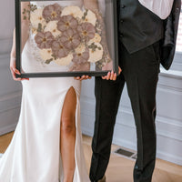 A happy couple stands holding their wedding bouquet preservation featuring pressed white roses and pressed purple roses with accents of pressed baby's breath and pressed eucalyptus. 