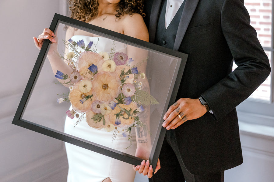 Holds hold a bouquet preservation piece featuring pressed florals and greenery while wearing wedding attire in a brightly lit room.