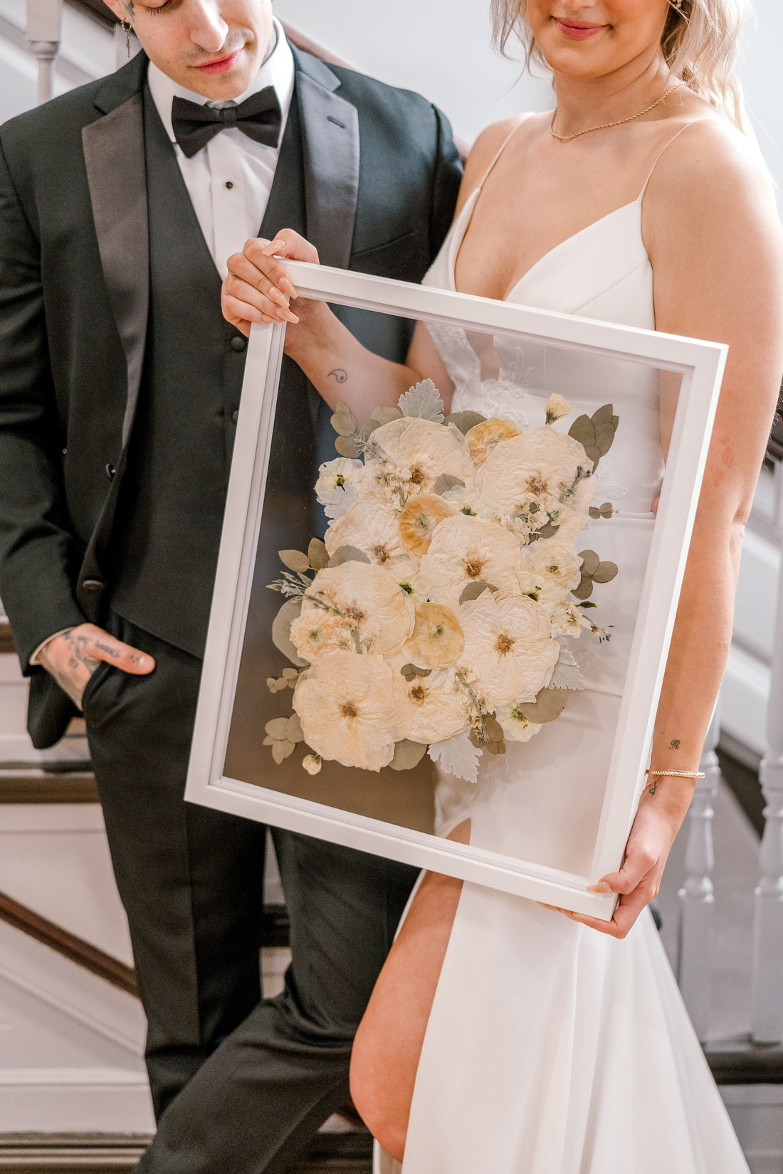A happy couple admiring their bouquet preservation piece featuring white pressed flowers and pressed greenery accents in a white wood floating frame. 