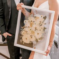 A happy couple admiring their bouquet preservation piece featuring white pressed flowers and pressed greenery accents in a white wood floating frame. 