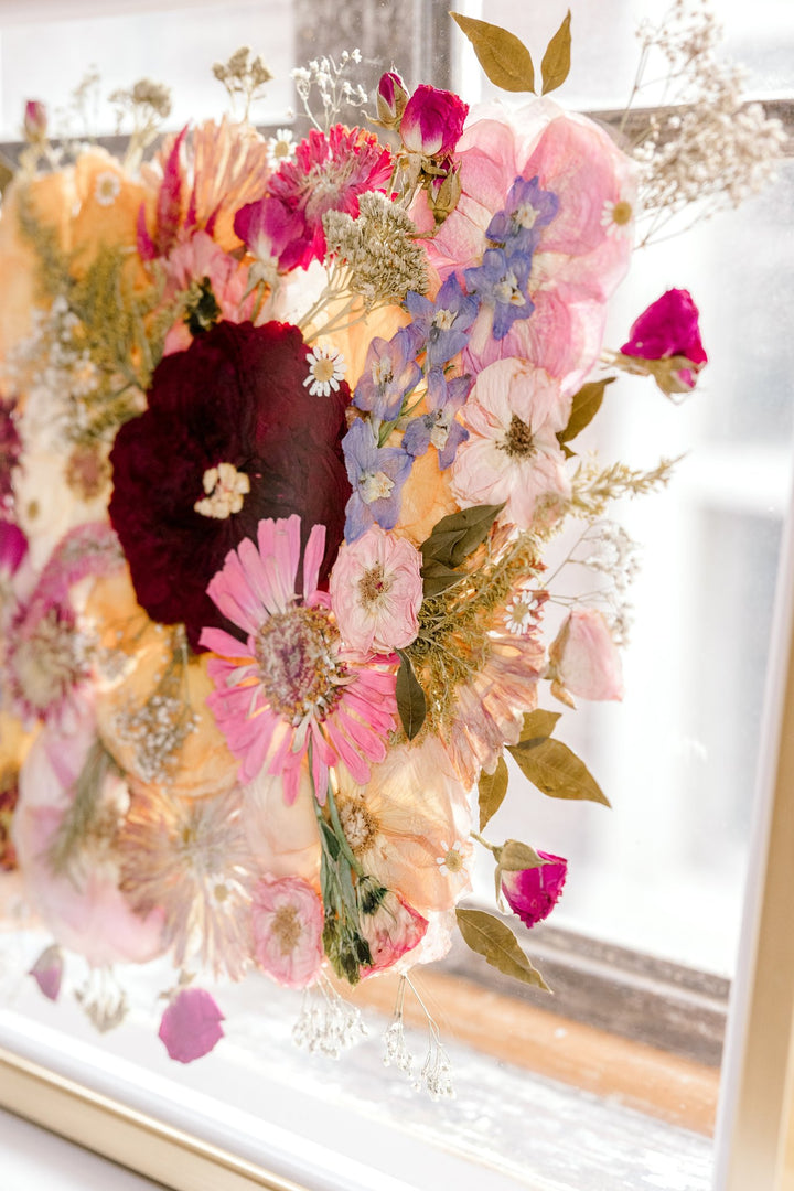 Brightly colored pressed florals in a gold floating frame with the light shining through. 