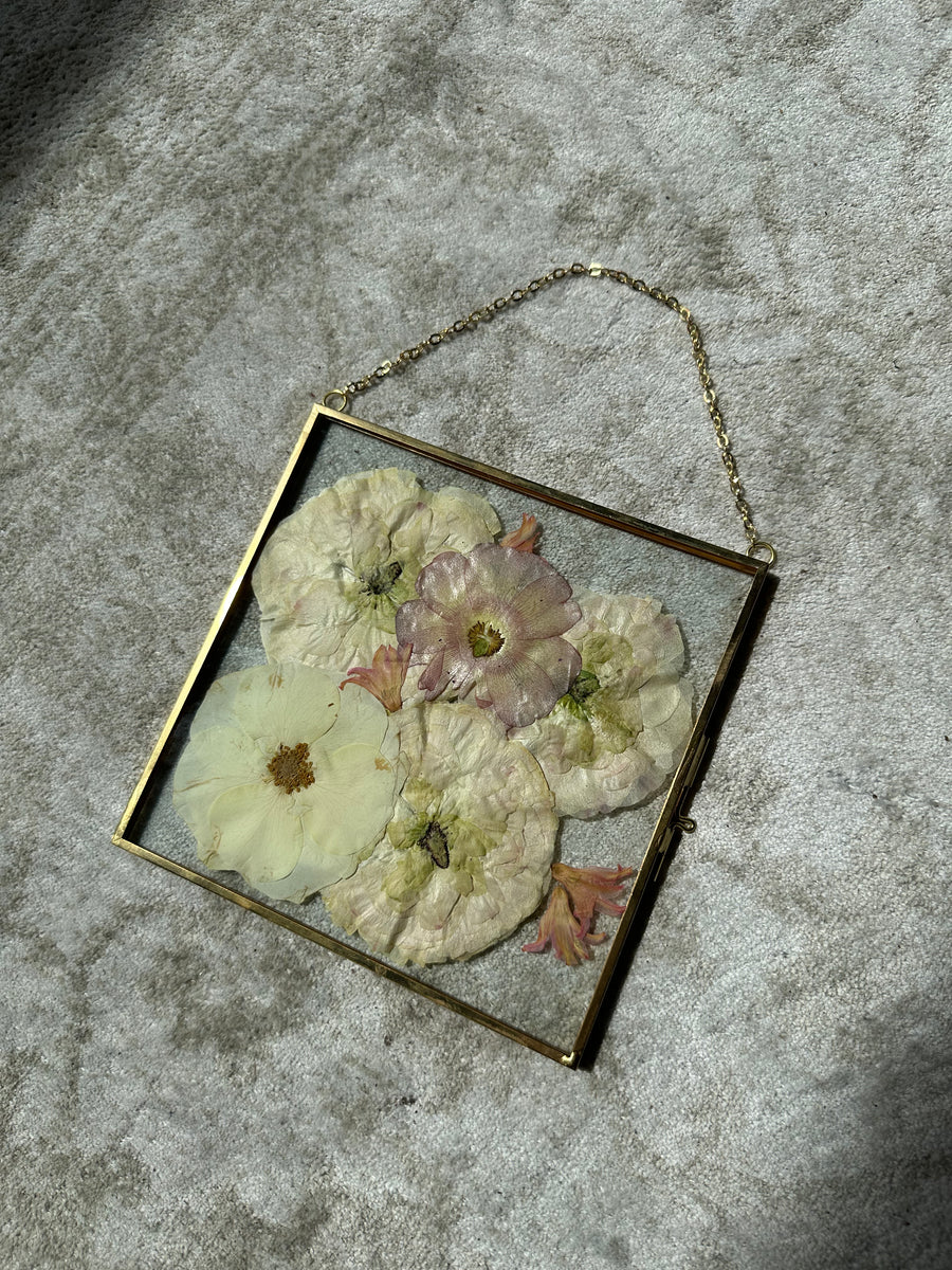 a small square gold hanging frame with pressed flowers against a white rug.