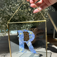 Gold hanging frame with pressed flower petals designed into the letter r held by a person.