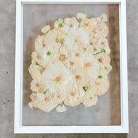 An all white ball bouquet pressed and preserved into a white frame against concrete.