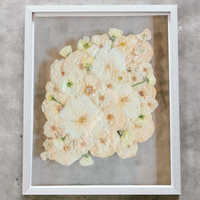 An all white ball bouquet pressed and preserved into a gray frame against concrete.