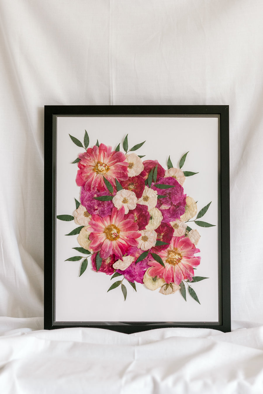 A bright pink and white bouquet is detailed by sprouts of greenery as they have been pressed and framed inside this bouquet preservation black wood frame with an archival grade background.
