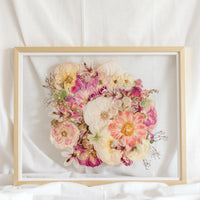 A bright pink and white pressed flower arrangement from a wedding bouquet is centered inside this gold wood frame.