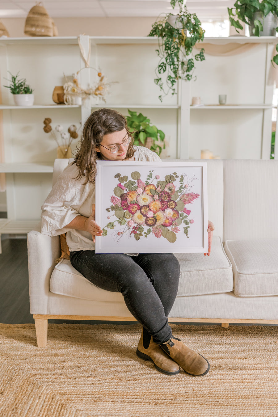A woman sits holding her bouquet preservation she received from the Pressed Bouquet Shop. It features pink and white pressed flowers with pressed greenery on a white background and in a white frame.