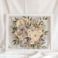 A white and light purple wedding bouquet turned into pressed flowers and displayed inside of a white wood preservation frame.