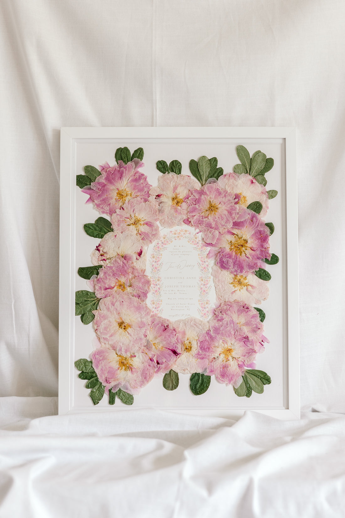 A bouquet of pressed pink peonies is displayed with a wedding invitation in a 16x20 white wood frame.