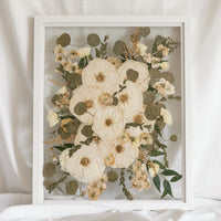 An all white and green pressed flower wedding bouquet displayed inside of a white wood glass floating frame.