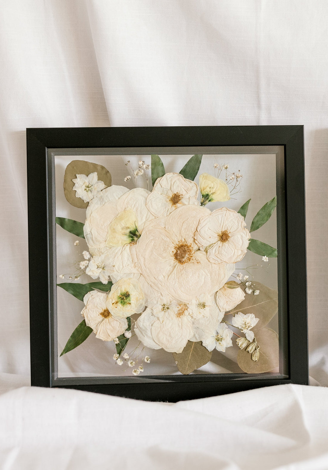 An all white and green bouquet preserved as a pressed flower frame in a 10x10 inch black wood square. 