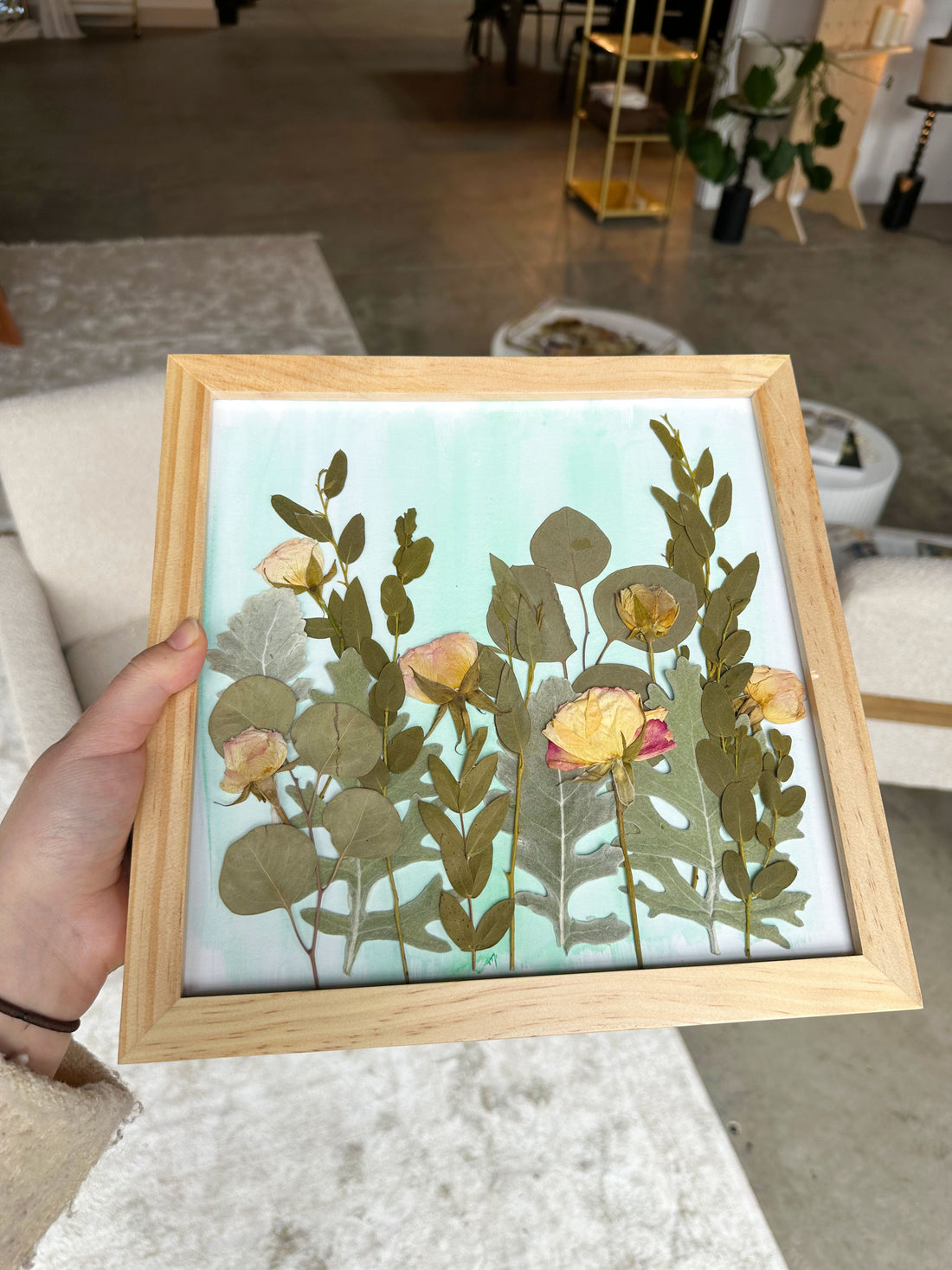 Person holding a tan colored frame that has pressed flowers in a field style design in their home.