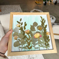 Person holding a tan colored frame that has pressed flowers in a field style design in their home.