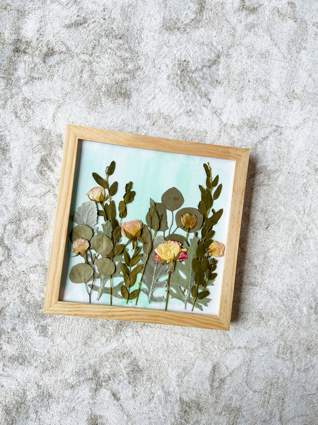 Small frame with pressed flowers in a field style design and water color background on top of a white carpet.