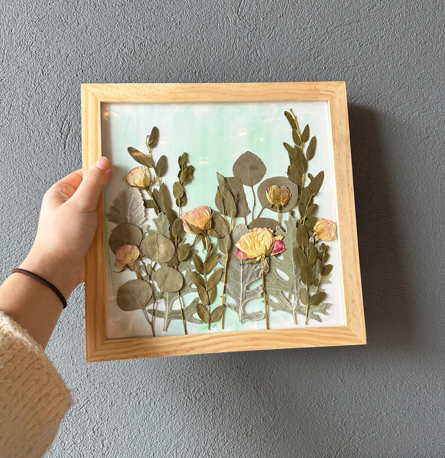 Person holding a natural colored frame encasing pressed flowers on a watercolor background in front of a grayish blue wall.