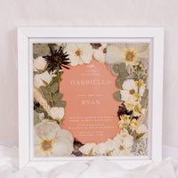 Beautifully preserved pressed flowers surround an orange-color wedding invitation in a square wood frame. 