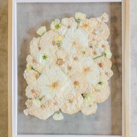 An all white ball bouquet pressed and preserved into a gold frame against concrete.