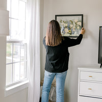 A woman stands in a bedroom hanging a frame filled with pressed flowers. 