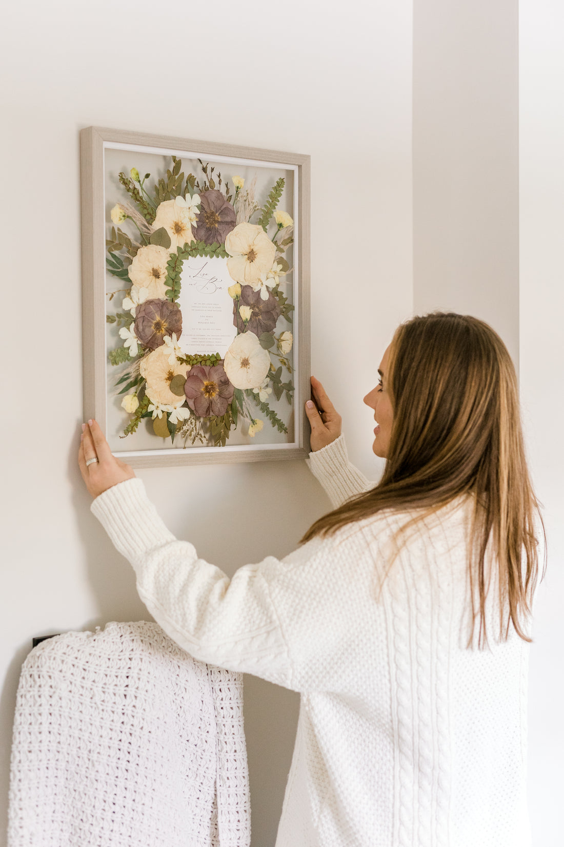 A woman hangs her pressed wedding bouquet on a wall above a blanket rack. The pressed flowers surround her wedding invitation that she smiles up at while hanging.
