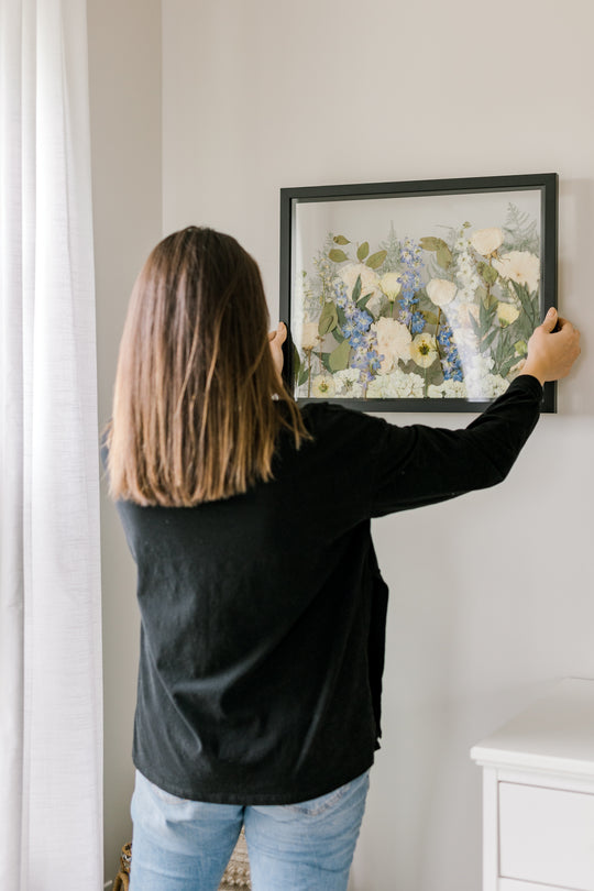 A pressed floral frame by Element Preservation, previously known as Pressed Bouquet Shop, being hung by a woman in their bedroom. 