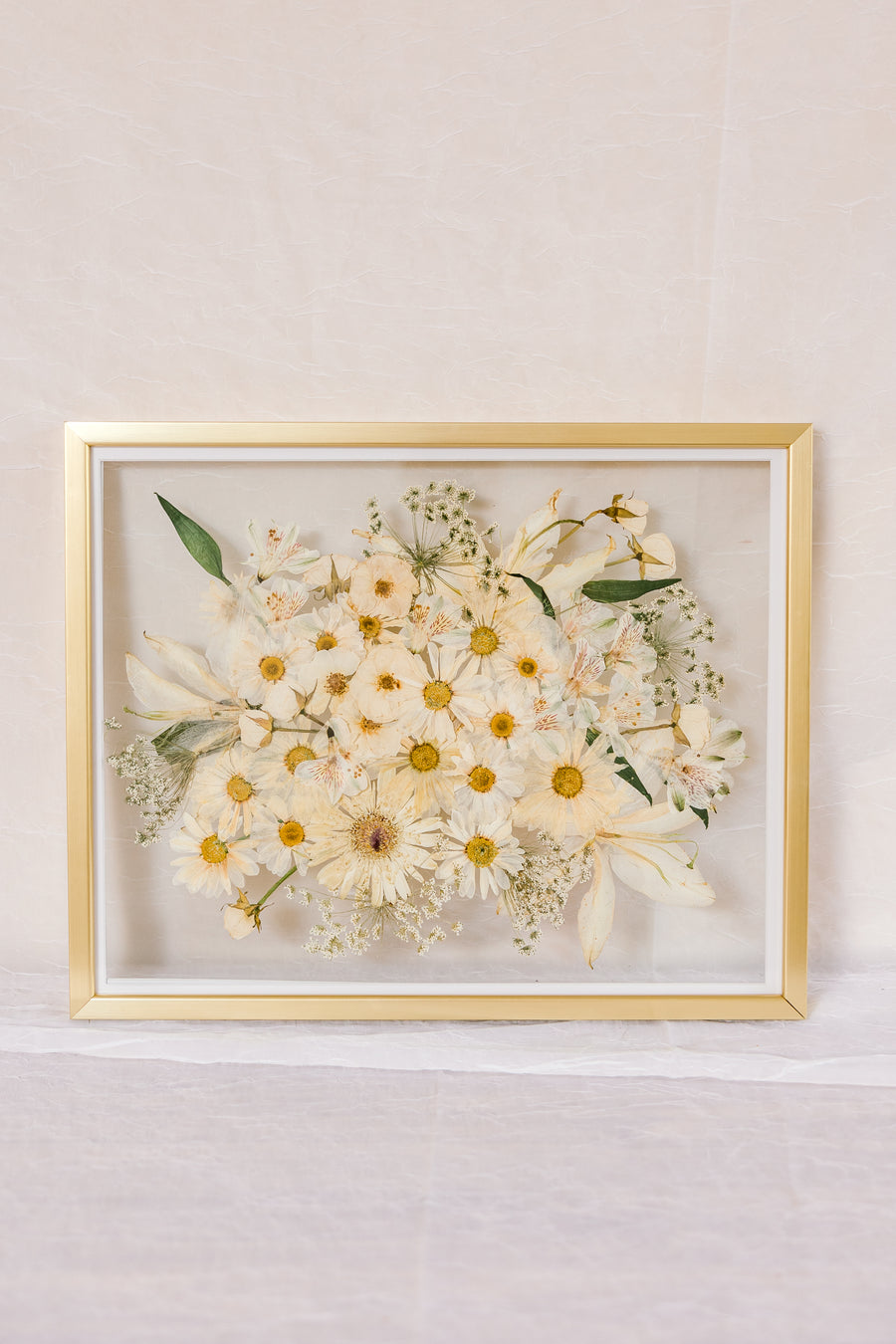 An all white bouquet of flowers pressed into a glass floating frame surrounded by gold wood.