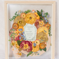 Orange and blue flowers surrounding a wedding invitation with ribbon and a wax seal, preserved in a glass floating frame. 